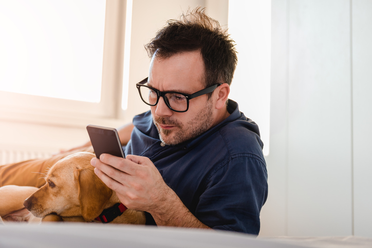 Man with glasses in blue shirt laying on the bed with the dog and using smart phone.
